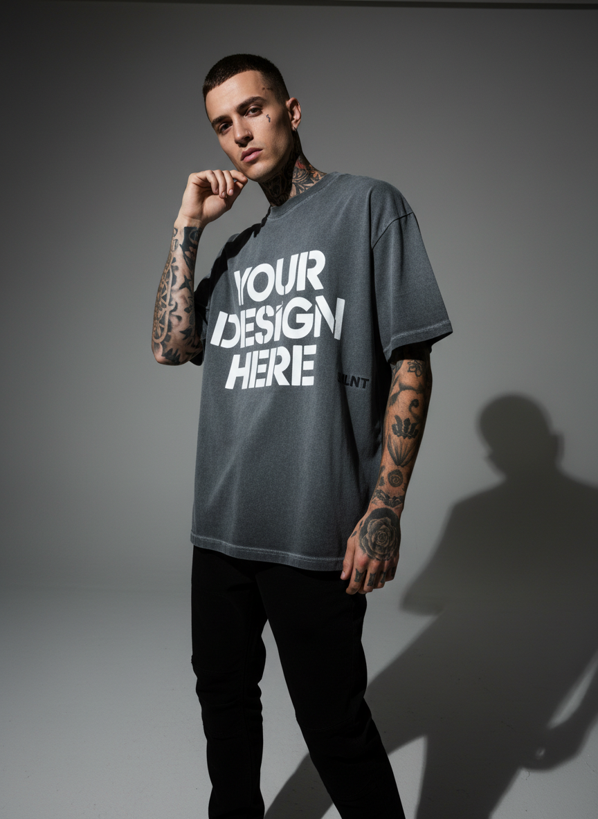 Tattooed male model in a grey graphic oversized tee and black pants, studio shot with dramatic lighting.